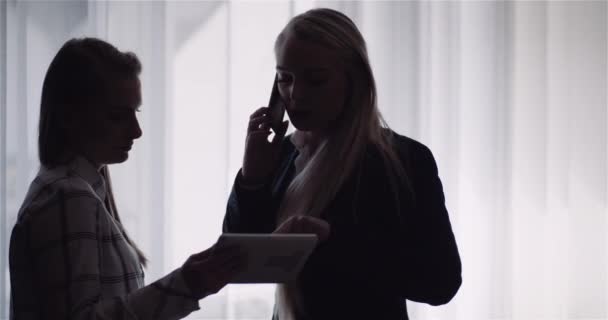 Две женщины обсуждают планшетный дисплей, в то время как одна женщина также разговаривает на смартфоне — стоковое видео