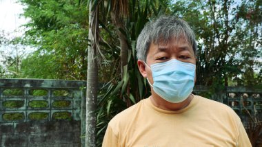 Asyalı orta yaşlı bir adam yüzüne koruyucu bir maske takıyor ve arka planda yeşil bitkiler var. Toz gazlarının zararlı etkilerinden korunuyor.