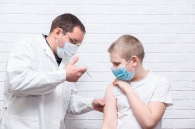 The smiling boy is looking away while his doctor holding a syringe next to his arm. Pediatrician makes vaccination to cute Caucasian boy. Boy in medical face mask getting flu shot by doctor