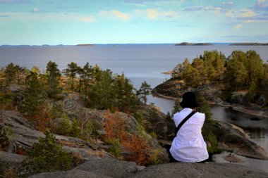 girl tourist in a white shirt and black panama sits admiring the views of the skerries, the rocky islands of Lake Ladoga on a summer day clipart
