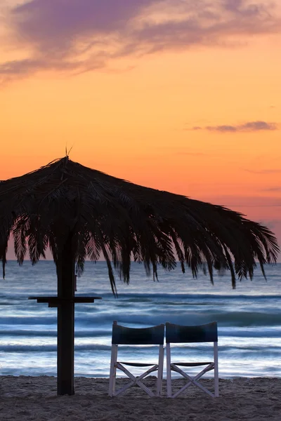 秸秆大伞，在海上日落场景的两个空沙滩椅。. — 图库照片