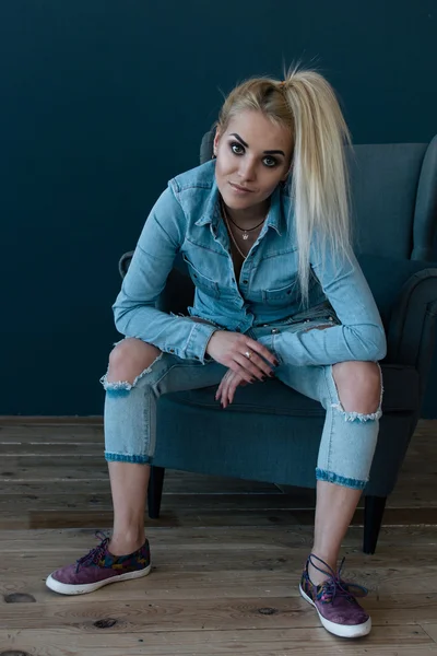Mladá sexy blondýna v modré košili a džínách, sedí v křesle — Stock fotografie