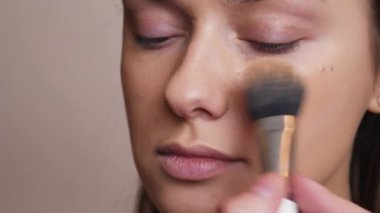 Makyöz kadın yüzüne bej rengi sıvı bir temel uyguluyor.