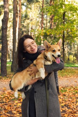 Gri montlu bir kadın sonbahar parkında köpeğiyle yürüyor.