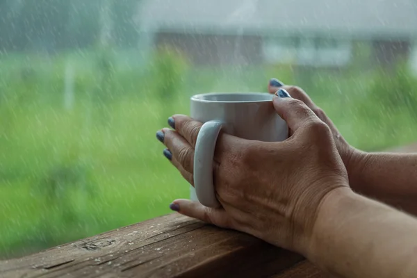 Manos femeninas sosteniendo una taza de café en el día lluvioso —  Fotos de Stock