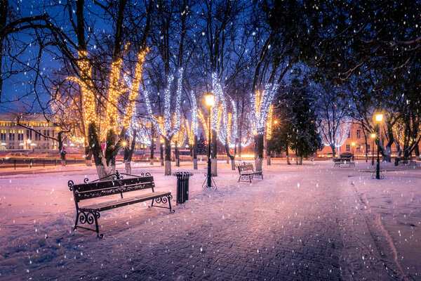 Winter night park with lanterns, benches and Christmas decorations in heavy snowfall.
