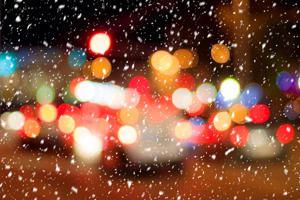 Falling snow against the background of blurry lights of headlights of cars and the night city. Winter snowfall concept. Abstract bright bokeh.