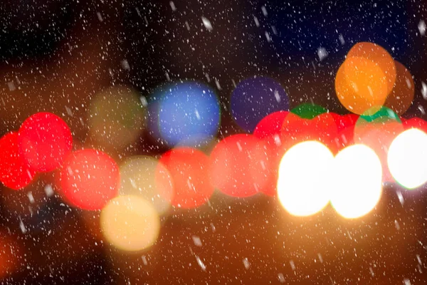 Falling snow against the background of blurry lights of headlights of cars and the night city. Winter snowfall concept. Abstract bright bokeh.