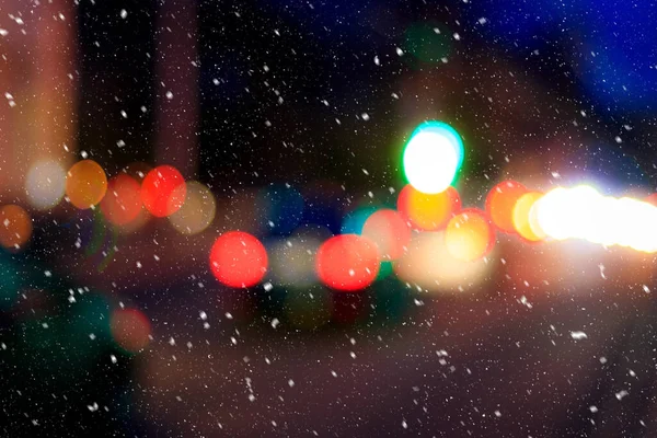 Falling snow against the background of blurry lights of headlights of cars and the night city. Winter snowfall concept. Abstract bright bokeh.