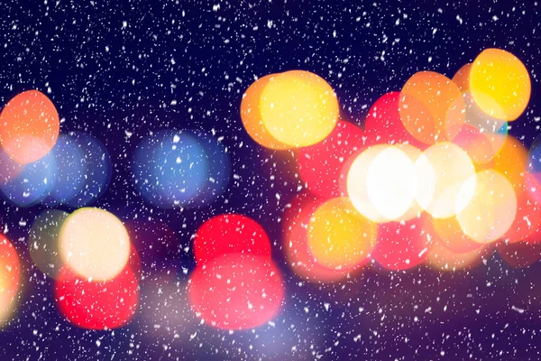 Falling snow against the background of blurry lights of headlights of cars and the night city. Winter snowfall concept. Abstract bright bokeh.