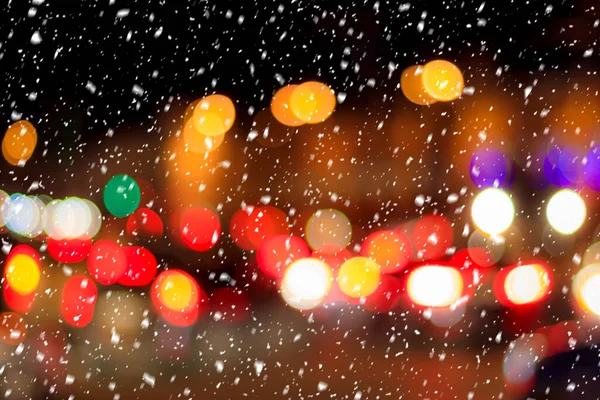 Falling snow against the background of blurry lights of headlights of cars and the night city. Winter snowfall concept. Abstract bright bokeh.