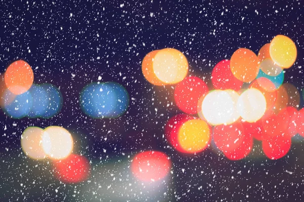 Falling snow against the background of blurry lights of headlights of cars and the night city. Winter snowfall concept. Abstract bright bokeh.