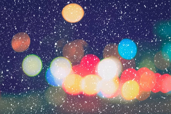 Falling snow against the background of blurry lights of headlights of cars and the night city. Winter snowfall concept. Abstract bright bokeh.