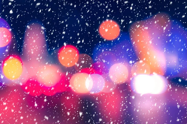 Falling snow against the background of blurry lights of headlights of cars and the night city. Winter snowfall concept. Abstract bright bokeh.