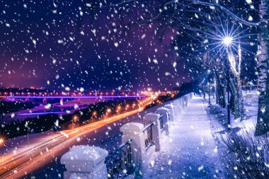 Geceleri bir kış parkında fenerlerle kar yağışı, araba hareketleriyle yola bakmak, kaldırımlar ve karla kaplı ağaçlar..