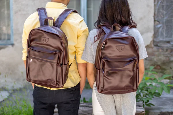 Back of woman and man with brown leather backpack. Man and woman holding hands and presenting leather bags. Unisex bag for sale.