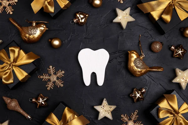 White tooth with golden decorations and gift boxes on black background. Dentist Merry Christmas and New Year concept. Top view, flat lay.