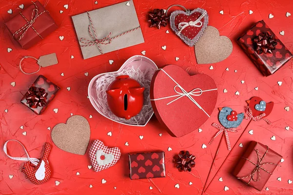 Piggy bank in a heart shape gift box surrounded by hearts, gifts and other love things on red background. Saving money for St. Valentines Day. Top down composition.