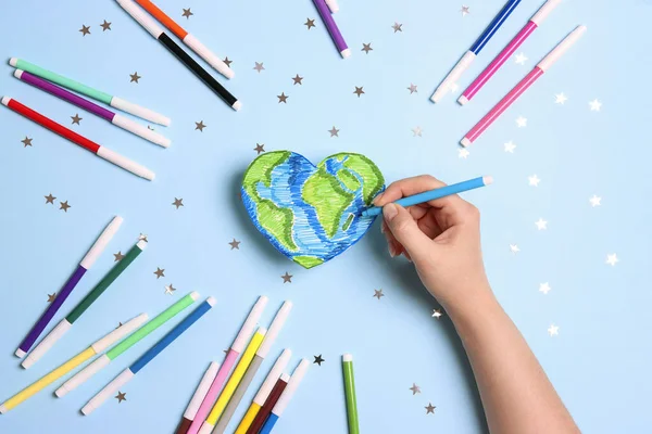 Female hand draws planet Earth  in the shape of a heart with multicolored felt-tip pens on on a blue background. Science, ecology, environmental protection. World Earth Day concept.