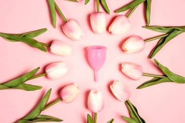 Pembe arka planda lalelerle çevrili regl bardağı. Adet görme, koruma, kadın sağlığı..