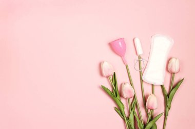 Farklı tipte kadın hijyen ürünleri. Tamponlar; adet bardağı ve pembe arka plandaki lale çiçekleri gibi hijyenik pedler. Kopyalama alanı olan üst görünüm.