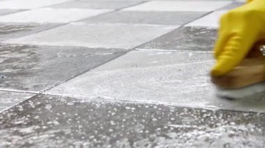 Sarı lastik eldiven takan eller döşeme temizleyicisiyle yerleri fırçalamak için ahşap bir zemin temizleyicisi kullanıyor..