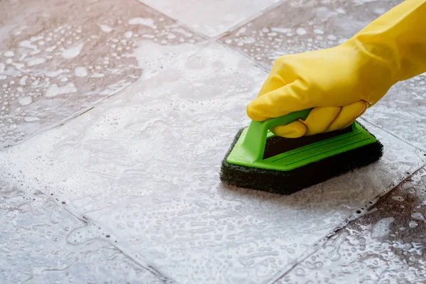 黄色のゴム手袋を身に着けている人間の手は 床掃除機でタイルの床をこするために緑の色のプラスチック製の床スクラバーを使用しています — ストック写真