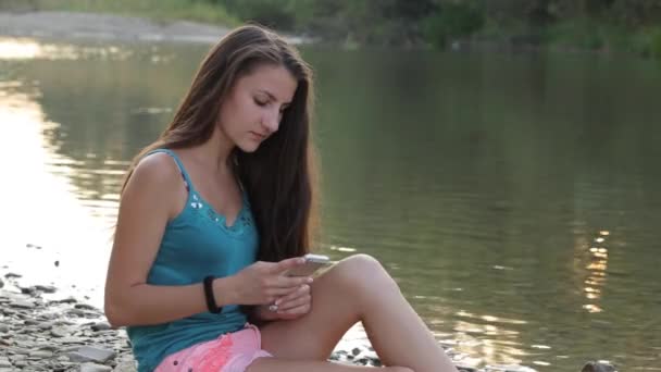 Девушка, сидящая на берегу реки с печатанием телефона — стоковое видео