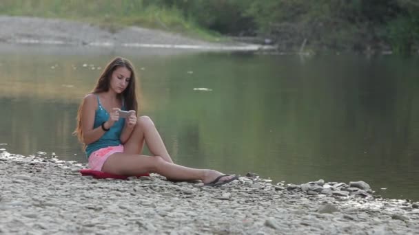 Девушка сидит на берегу реки с телефоном играет в игру — стоковое видео