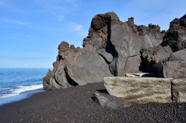 Siyah kum ve kayalarla kaplı bir plajda volkanik katılaşmış lavların yakın çekimi. Sol tarafta denizi görebilirsiniz ve arka planda mavi bir gökyüzü var.