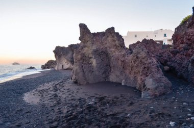 Siyah kumsalda lav kayaları var. Pencerelerinde mavi panjurlu kar beyazı bir ev var. Gün batımı ve ufukta bir gemi var. .