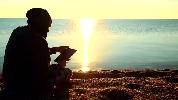 En kille med en tablett som sitter på grus stranden framför havet — Stockvideo