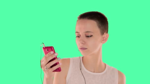 Menina careca bonita, com cabelo curto morena na tela verde, ouvindo música, telefone celular, fones de ouvido — Vídeo de Stock