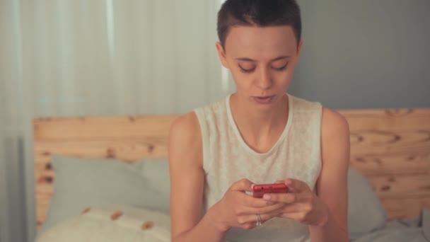 Hermosa chica calva, una chica con pelo corto morena sentada en la cama riendo mientras revisa su teléfono celular — Vídeos de Stock