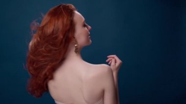 Uzun kıvırcık kızıl saçlı güzel, modaya uygun bir kız. Stüdyoda mavi arka planda bir kız var. Reklamlar, saç ürünleri, güzellik salonu, kozmetik ürünleri, kıyafetler. Moda, butik.
