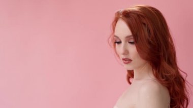 Uzun kıvırcık kızıl saçlı güzel, modaya uygun bir kız. Stüdyoda pembe arka planda bir kız var. Reklamlar, saç ürünleri, güzellik salonu, kozmetik ürünleri, kıyafetler. Moda, butik. Pembe..