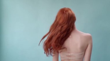 Uzun kıvırcık kızıl saçlı güzel, modaya uygun bir kız. Stüdyoda mavi arka planda bir kız var. Reklamlar, saç ürünleri, güzellik salonu, kozmetik ürünleri, kıyafetler. Moda, butik.