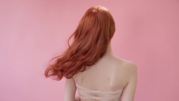 漂亮的时髦姑娘,一头长长的卷曲的红头发.一个粉红背景的女孩在工作室里广告，理发产品，美容院，化妆品，服装。时尚，精品。粉红. — 图库视频影像