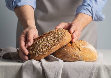 Önlüklü fırıncı gri arka planda taze pişmiş ekmeği masanın üzerinde tutuyor.