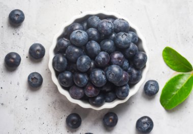 Beyaz bir kasede olgun yaban mersinleri ve gri beton bir zemine serpilmiş yaprak manzaralı.
