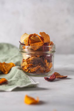 Kavanozda patates, pancar ve havuçtan yapılmış sebze cipsleri ve bir peçeteyle beton bir arka plana yerleştirilmiş.