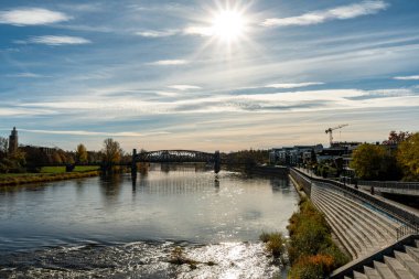 Sakin bir nehir üzerinde güneşli bir sonbahar manzarası çelik köprüsü ve nehir kenarındaki gezinti güvertesi. Yansıtıcı su ve yumuşak öğleden sonra ışığı ile geniş yatay bir formatta yakalanır..