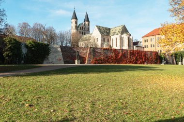 Magdeburg 'daki Meryem Ana Manastırı' nda açık bir sonbahar günü. Tarihi taş duvarlar ve renkli yapraklar üzerinde sıcak bir ışık. Önplanda açık yeşil alan ile geniş yatay bir kompozisyonda yakalanır.