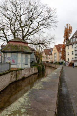 Sessiz, eski bir kasaba kanalı, yumuşak gri gün ışığı altında dikey bir kompozisyonda yakalanmış, yarım keresteli bir pavyonun ve sonbahar evlerinin yanından akar..
