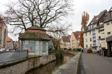 Sakin, eski bir kasaba kanalı, yumuşak gri gün ışığında yakalanan ve temiz kentsel alanla çerçevelenmiş, küçük, yarı keresteli bir pavyonun ve konut binalarının yanından geçiyor..