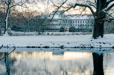 Sakin bir nehir, bir şehir parkının ağaçlarını ve kıyılarını kaplayan soğuk sabah ışığını yansıtır. Kış sahnesi, klasik bir yatay kompozisyonda sessiz ve dengeli görünüyor..