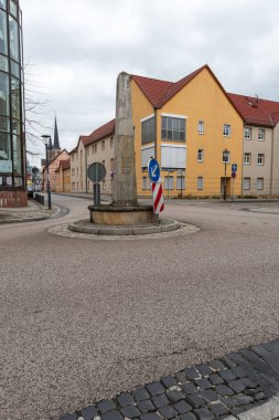 A historic stone distance marker stands on a small traffic island at an urban intersection. In the background a church tower rises above the rooftops of the small town, captured in vertical composition.