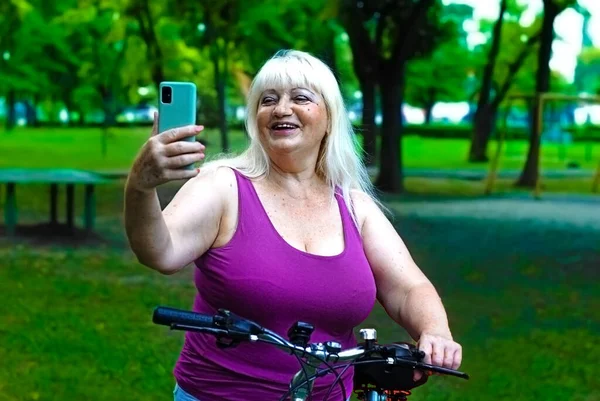 An elderly blonde woman is engaged in a bike ride in the park and takes a selfie. An elderly woman communicates by video call via a smartphone while walking a bicycle in the park.