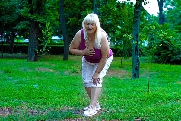 An elderly blonde woman felt unwell in the park. An elderly woman clutches her heart. An elderly woman is experiencing pain in the heart outdoors. Heart attack outdoors.