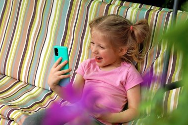 Küçük sarışın bir kız, yazlık bir salıncakta otururken akıllı bir telefonla video iletişimiyle iletişim kuruyor. Doğada küçük mutlu bir kız. Çocuk ve aletler.